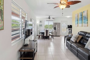 A spacious living area with a sofa, TV, dining table, chairs, ceiling fans, and artwork on the walls.