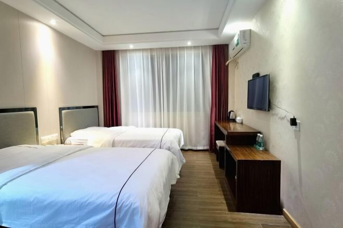A hotel room with two beds, a wooden desk, a television, and a large window with curtains.