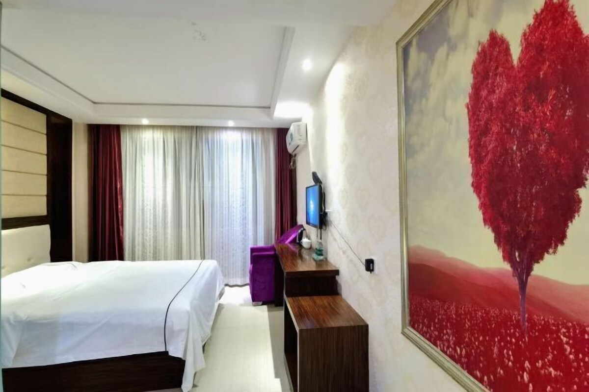 A hotel room with a bed, a desk, a TV, and a painting of a red tree.