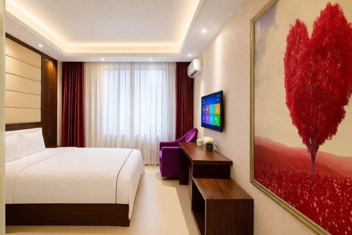 A hotel room with a bed, a desk, a chair, a TV, and a large painting of a red heart.