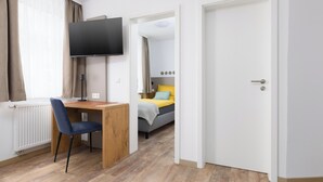 A hotel room with a wooden desk, a chair, a bed with yellow and blue bedding, a TV mounted on the wall, and a white door. - Gasthaus zur Traube (Uffenheim)