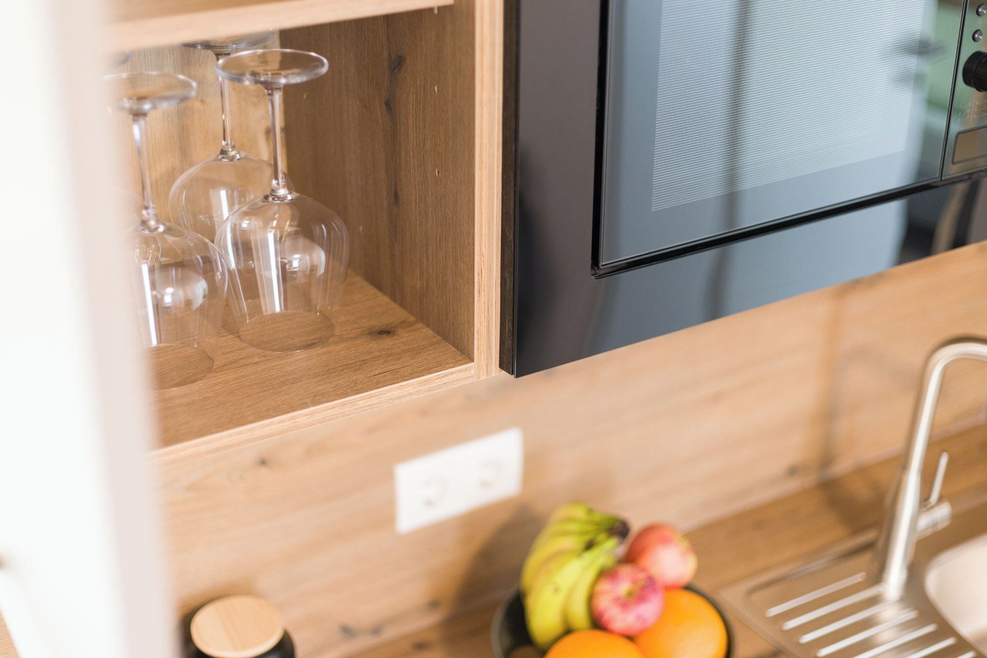 Une cuisine avec une étagère en bois où sont posés des verres à vin, un panier de fruits contenant des bananes, des pommes et des oranges, et un four à micro-ondes noir.