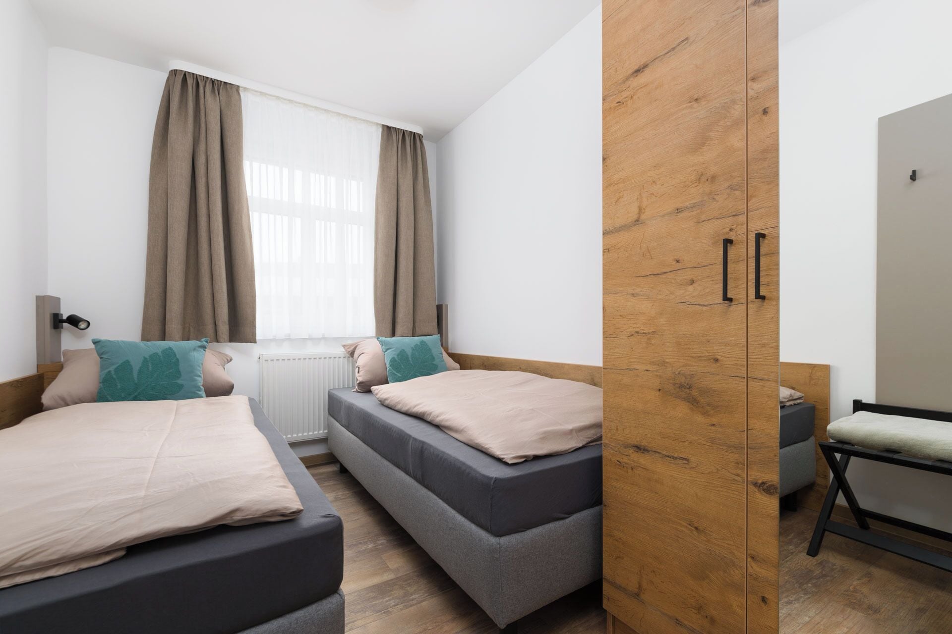 A modern hotel room with two beds, a wooden wardrobe, and a small seating area.