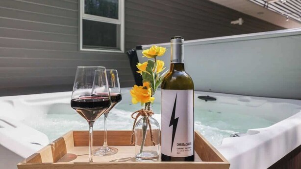 Ein Holztablett mit einer Flasche Charles & Charles Cabernet Sauvignon 2021, zwei Rotweingläsern und einer Vase mit gelben Blumen, platziert vor einem Whirlpool.