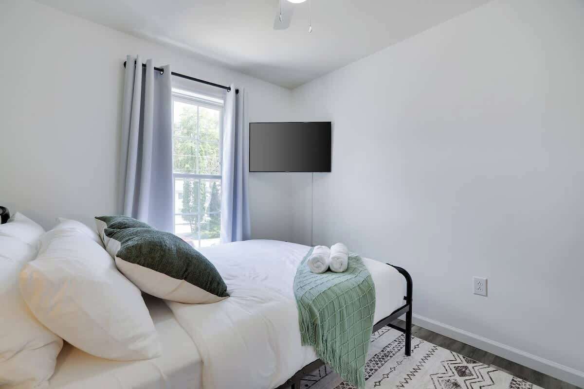 Ein Schlafzimmer mit Bett, Flachbildfernseher an der Wand, einem Fenster mit Vorhängen und einem gemusterten Teppich.