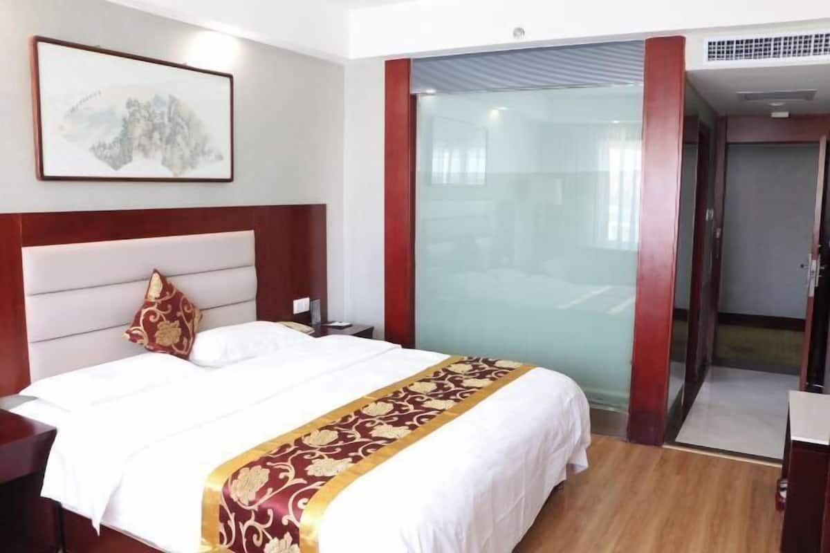 A hotel room with a large bed, a nightstand, a bedside table, a glass partition, and a door leading to another room.