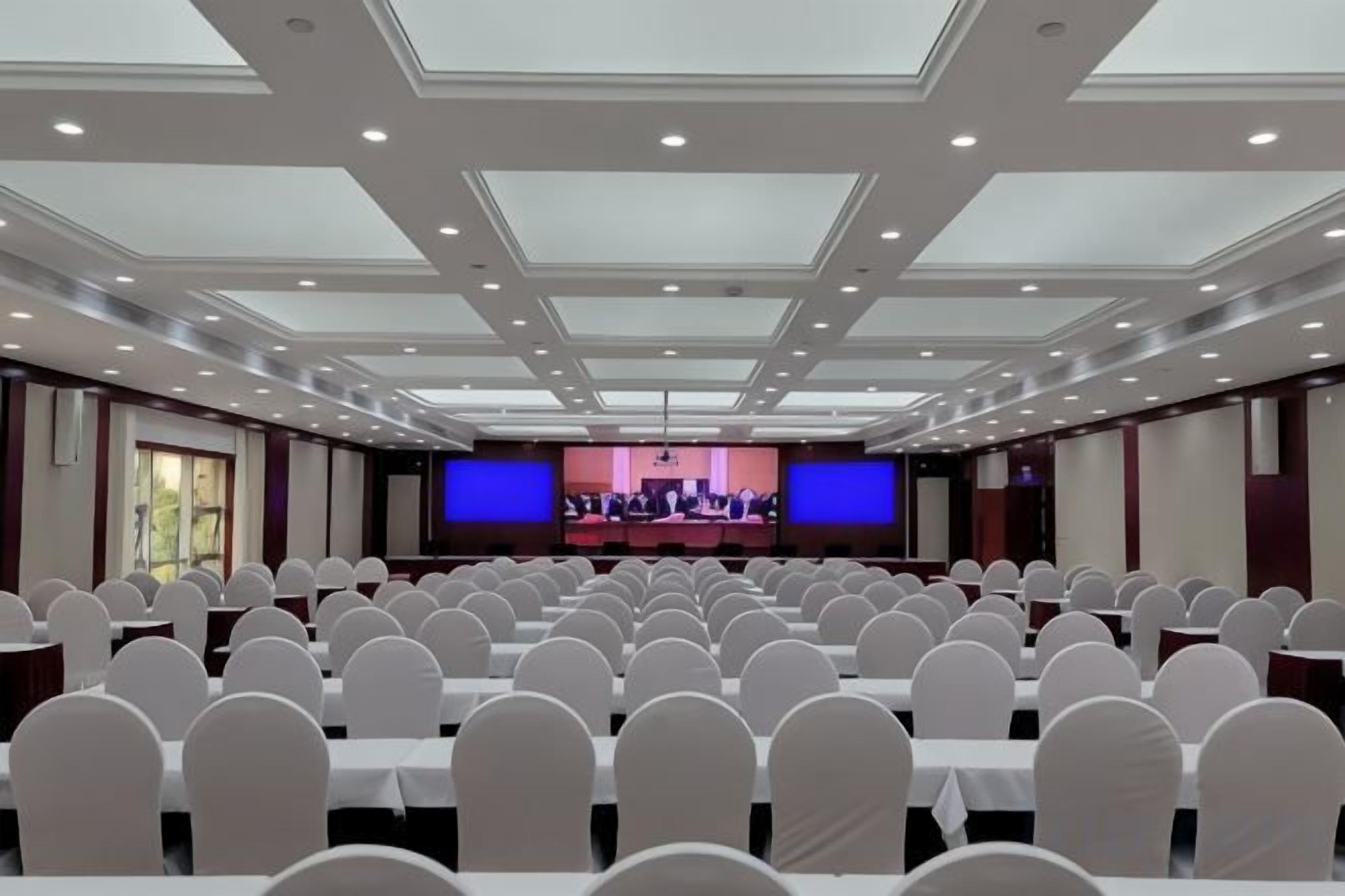A conference room with rows of tables and chairs, a stage with a podium, and a large screen.