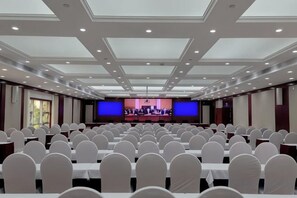 Une salle de conférence avec des rangées de tables et de chaises, une scène avec un pupitre et un grand écran.