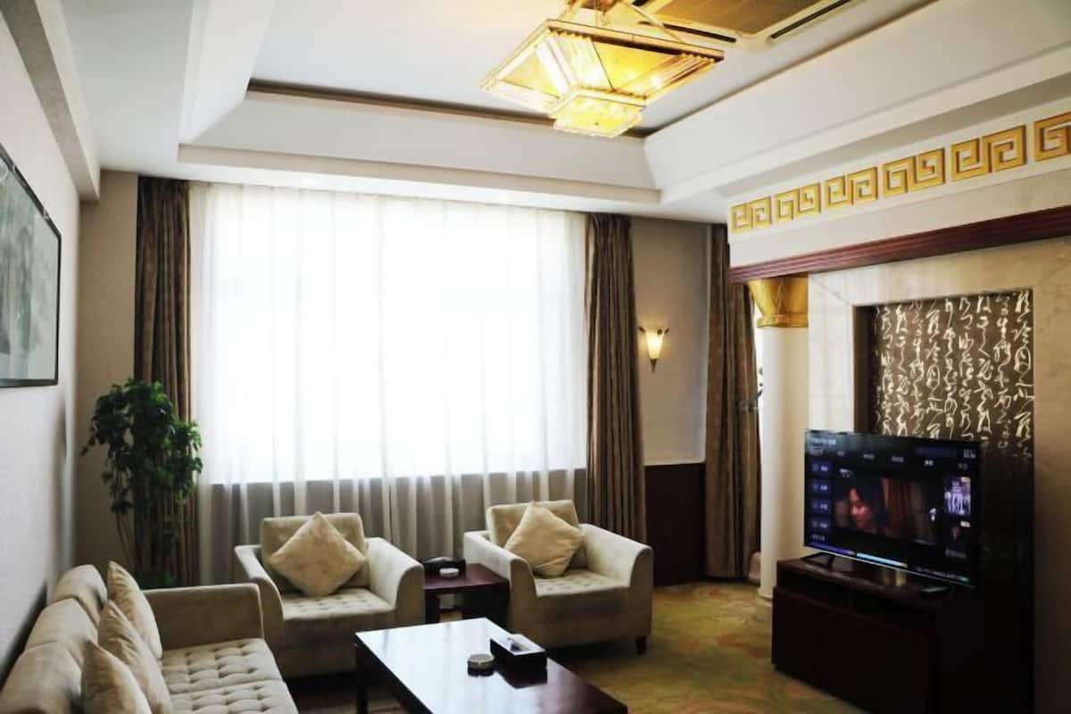 A hotel room with a sofa, two armchairs, a coffee table, a TV, and a large window with curtains.