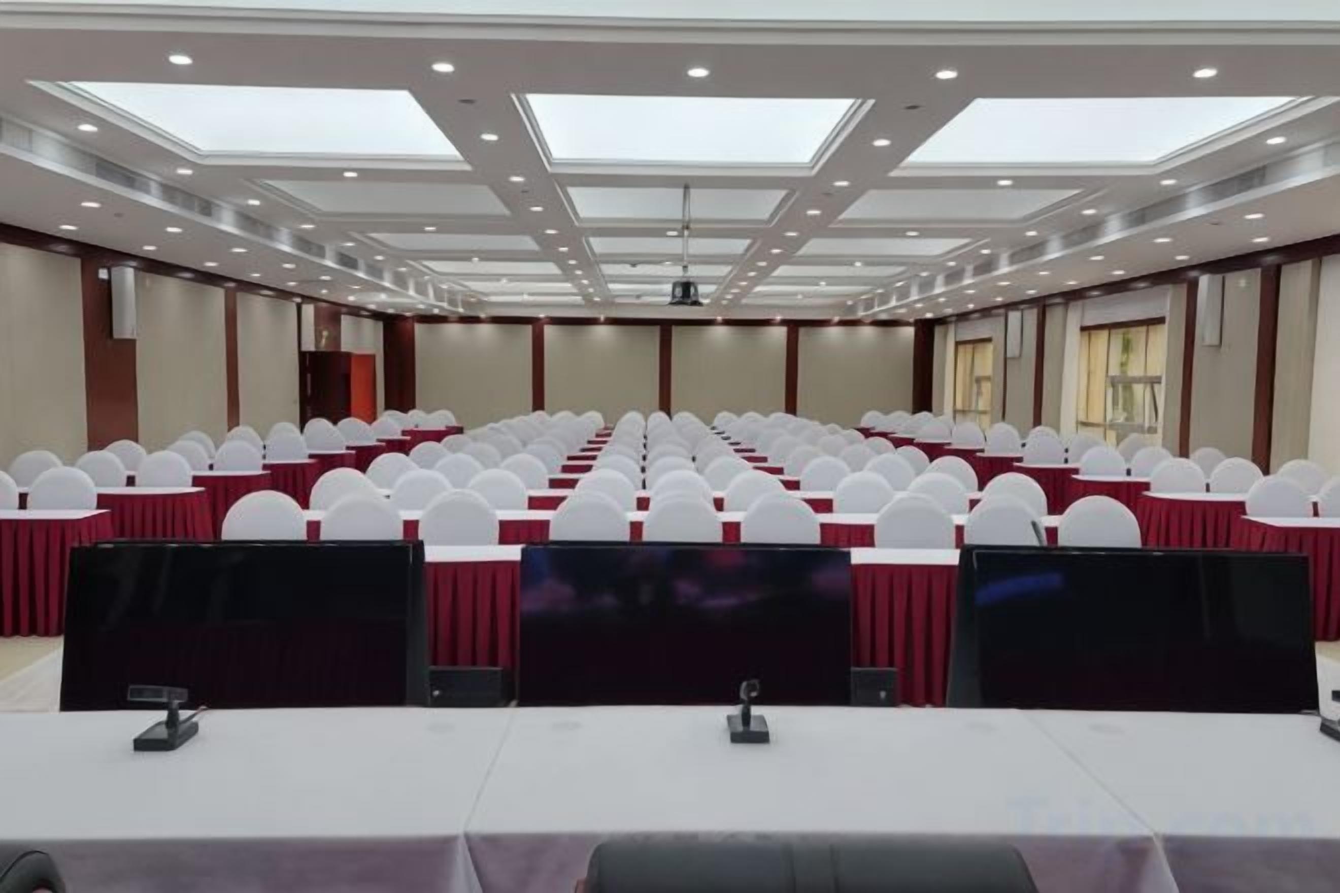 Un salón de conferencias con filas de sillas blancas, una mesa larga con mantel rojo y un techo con tragaluces.