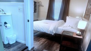 Une chambre d’hôtel comprenant un lit, une table de chevet avec une lampe, un petit tapis, une fenêtre avec des rideaux et une salle de bain avec des toilettes et une poubelle.