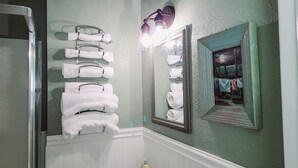 Une salle de bain équipée d’un porte-serviettes où sont rangées plusieurs serviettes blanches, d’un miroir avec un cadre décoratif et d’un espace douche.