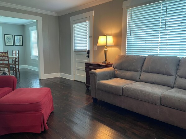 A living room with a grey sofa, a red ottoman, a wooden side table with a lamp, and a door with blinds. - The Redbird: 3 Bedroom, Pet-Friendly, Downtown, Fast WiFi, Porch (Douglas)