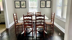 Une salle à manger avec une table blanche et six chaises en bois, de part et d’autre desquelles se trouvent deux fenêtres équipées de stores.