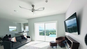 A modern living room with a grey sofa, a brown armchair, a flat-screen TV mounted on the wall, and a large sliding glass door leading to a balcony with a view of the ocean. - The Luxury Escape - Peaceful & Charming 3-bedroom villa w/ sea view (Vieux Fort)