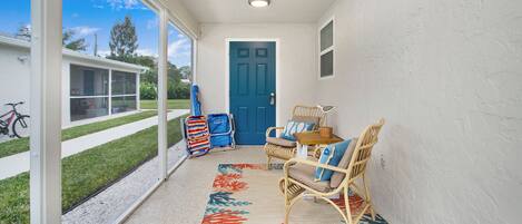 Une véranda couverte avec des fauteuils en osier, une petite table et un tapis coloré. Une porte bleue est visible au fond.