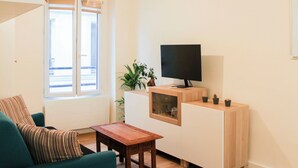 Ein Wohnzimmer mit einer Couch, einem kleinen Holztisch, einem Fernseher auf einer Kommode, einer Pflanze und einem Fenster mit Jalousien.