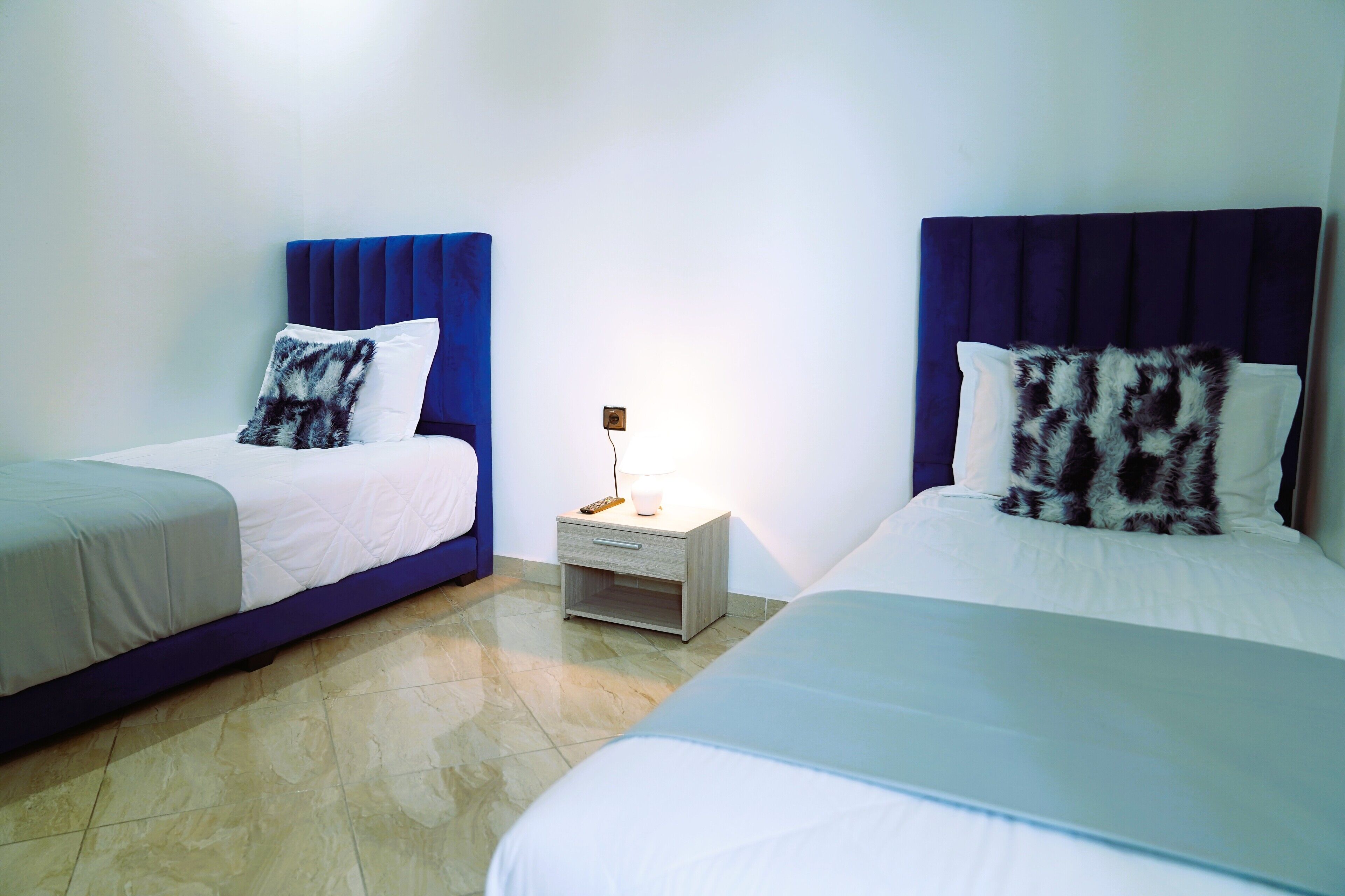Camera d'albergo con due letti, ognuno dotato di una testiera blu, un comodino con una lampada e un cuscino bianco con motivo nero e bianco.