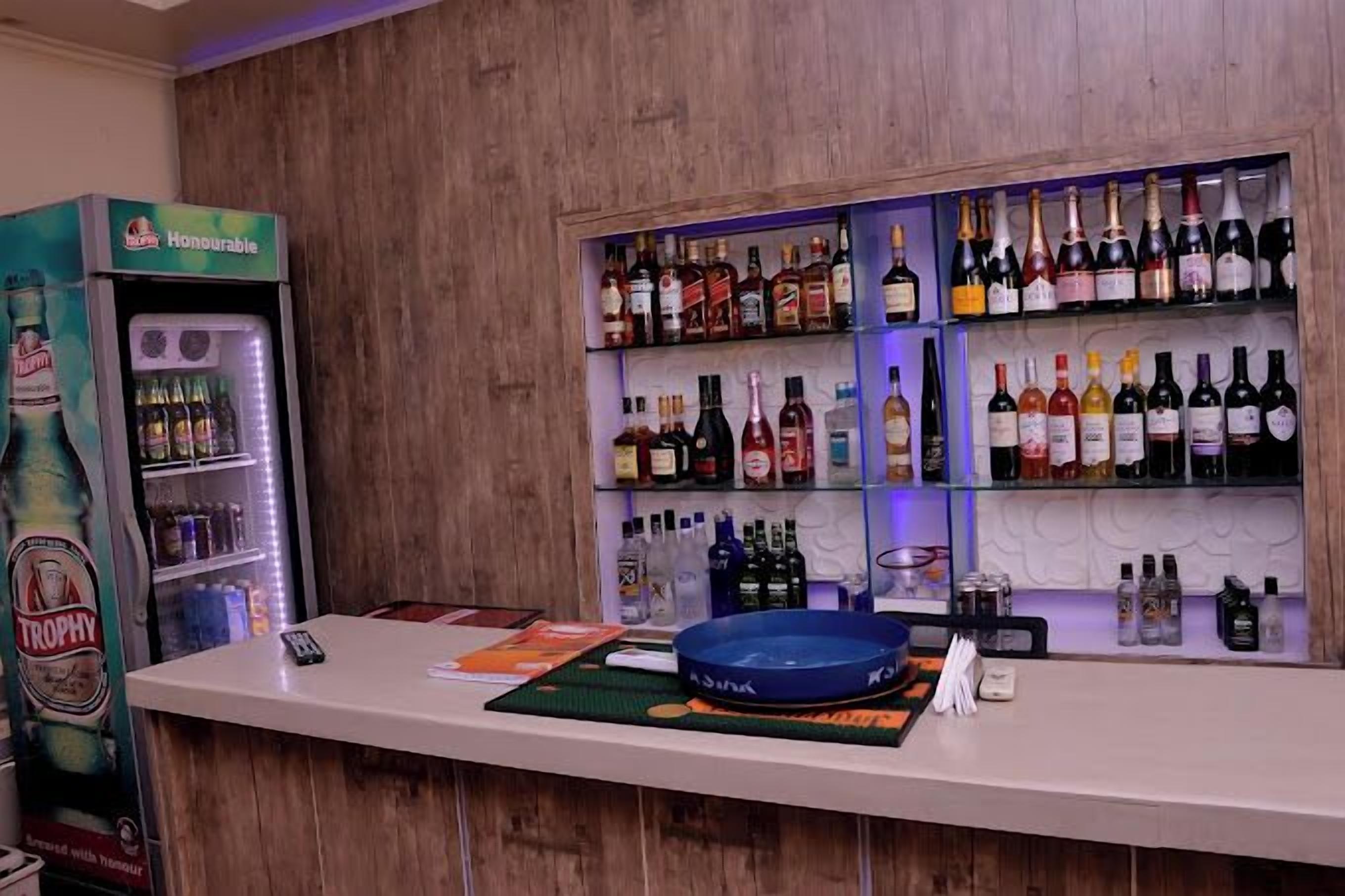 A bar area with shelves of various liquor bottles, a refrigerator with a branded beverage, and a counter with a phone and promotional materials.