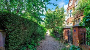 A narrow gravel path flanked by dense greenery and a brick wall with a metal gate. - Pass the Keys | Charming Victorian Terrace in centre of Cambridge (Cambridge)