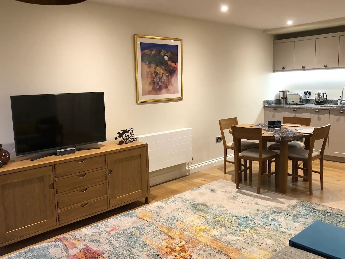 A living room with a TV, wooden cabinet, dining table with chairs, and a colorful rug.