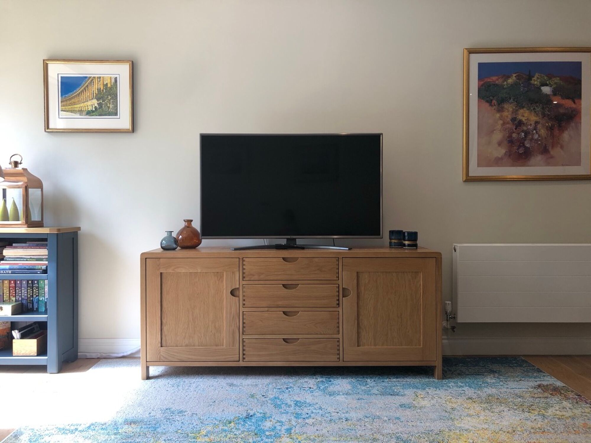 A living room with a flat-screen TV on a wooden cabinet, a bookshelf, and two framed pictures on the wall.