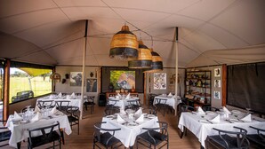 Un área de comedor con carpas, mesas redondas dispuestas para varios invitados, con manteles blancos y sillas negras.