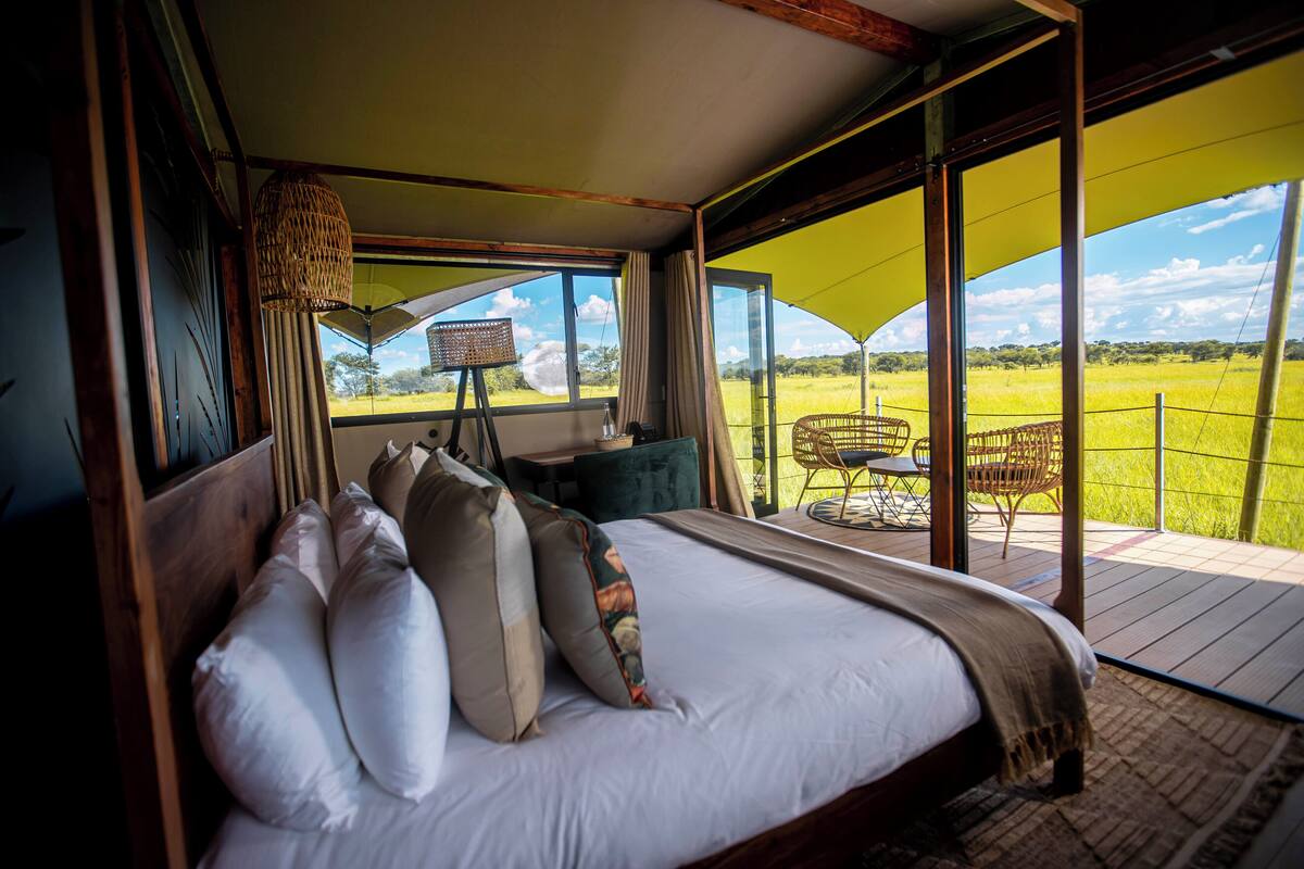 A bedroom with a large bed, a view of a grassy field, and a balcony with a chair.