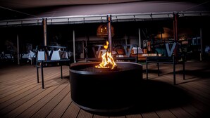 Una chimenea exterior circular con un banco de metal a cada lado, rodeada por una terraza de madera.