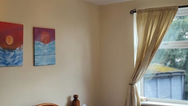 Un dormitorio con una cama, una mesita de noche, una lámpara de pared, dos cuadros y una ventana con cortinas.