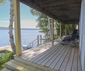 A wooden deck with a view of a lake, a covered area with chairs, and a wooden staircase leading to a dock.