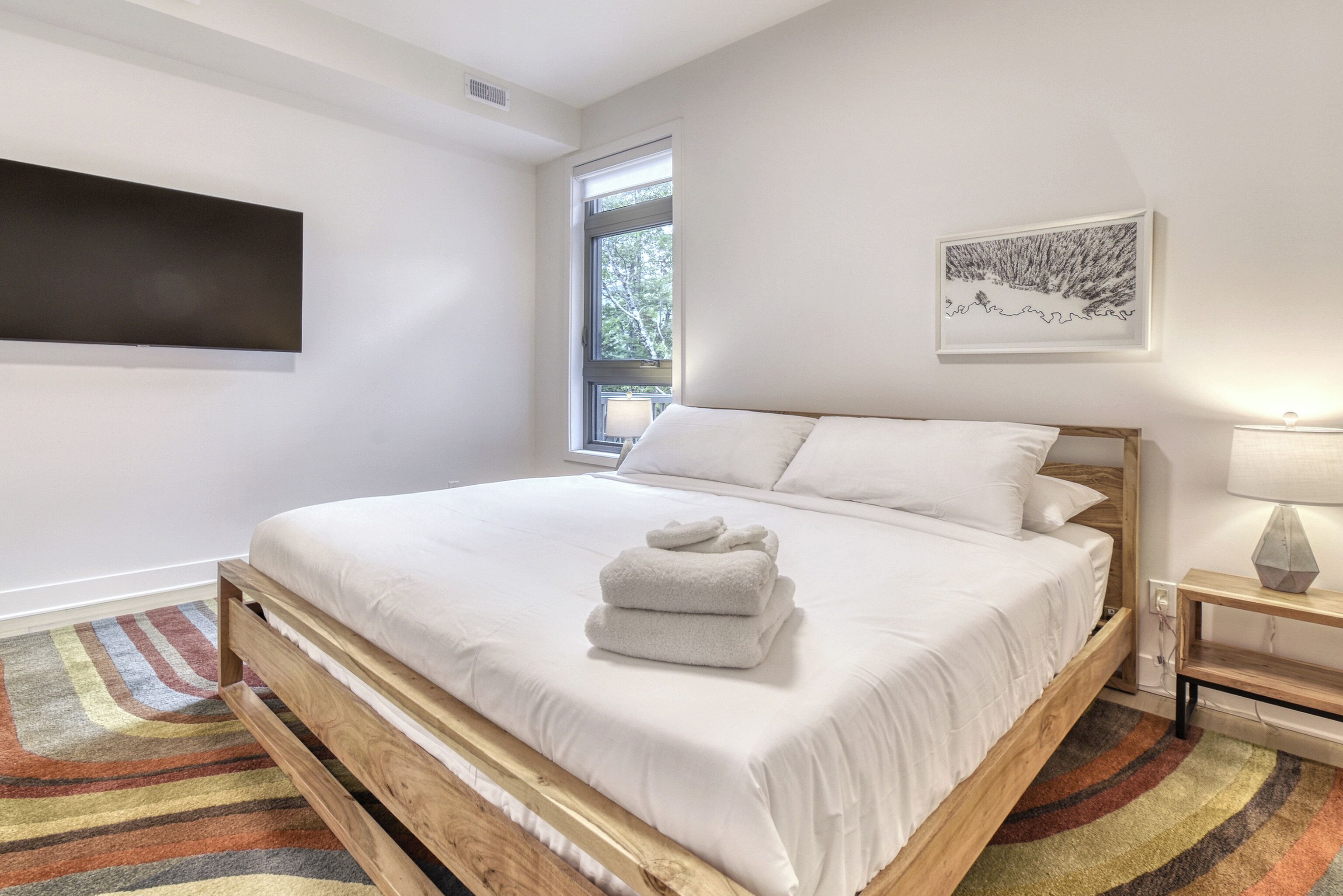 A neatly made bed with white linens and a folded towel, a wooden headboard, a colorful striped rug, a wall-mounted flat-screen TV, a window with a view of trees, and a framed picture on the wall.