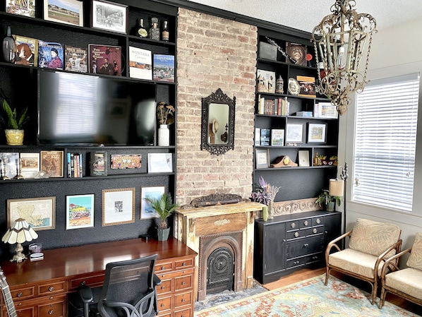 A cozy living room with a fireplace, a bookshelf filled with books and memorabilia, a desk with a chair, and a comfortable armchair.