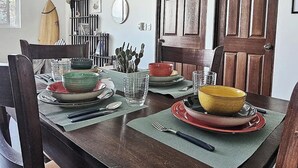 A dining area with a wooden table set for four, surrounded by chairs. There is a surfboard on the wall, a cactus plant, and a mirror.