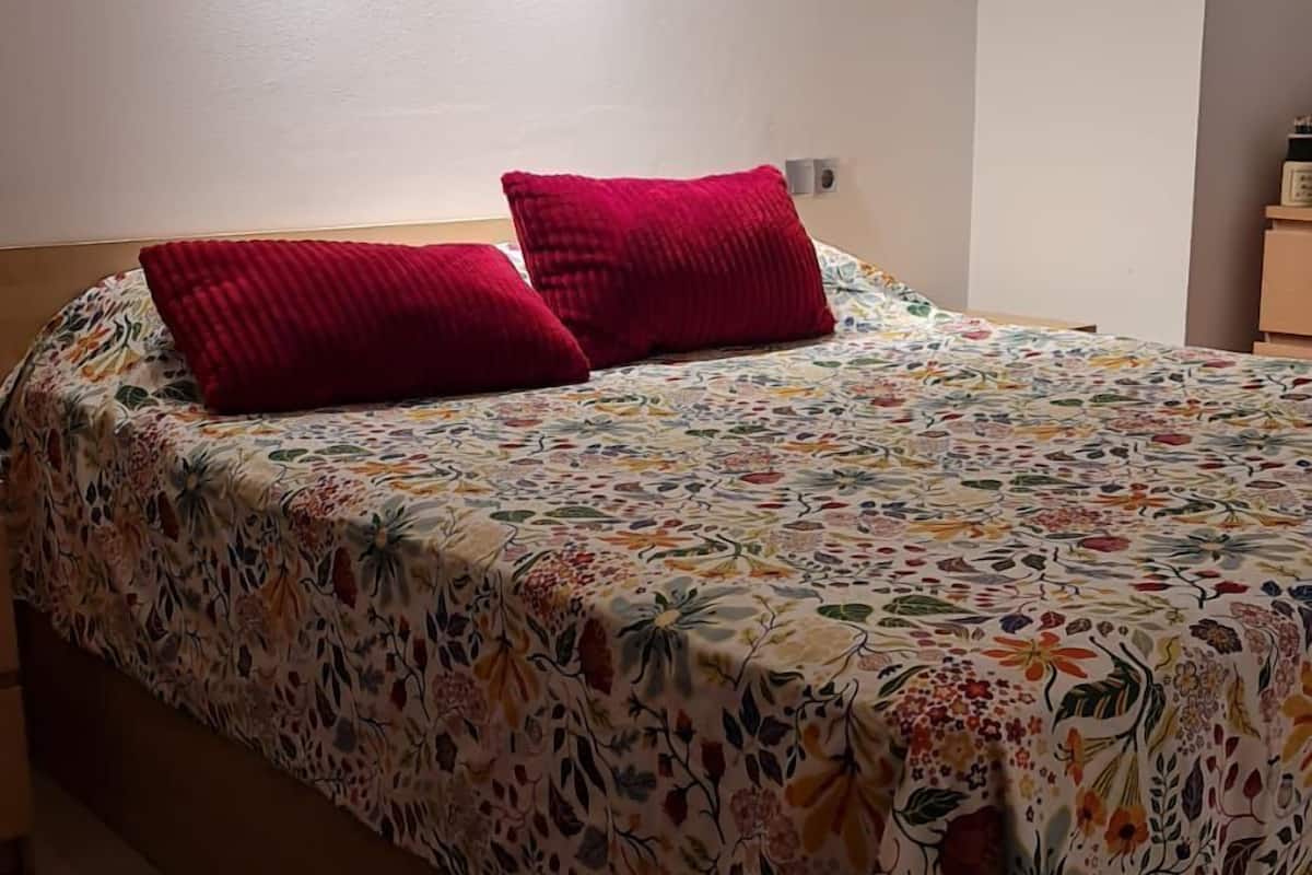 Una cama con colcha floral y dos almohadas rojas, cabecera de madera y lámpara de techo.