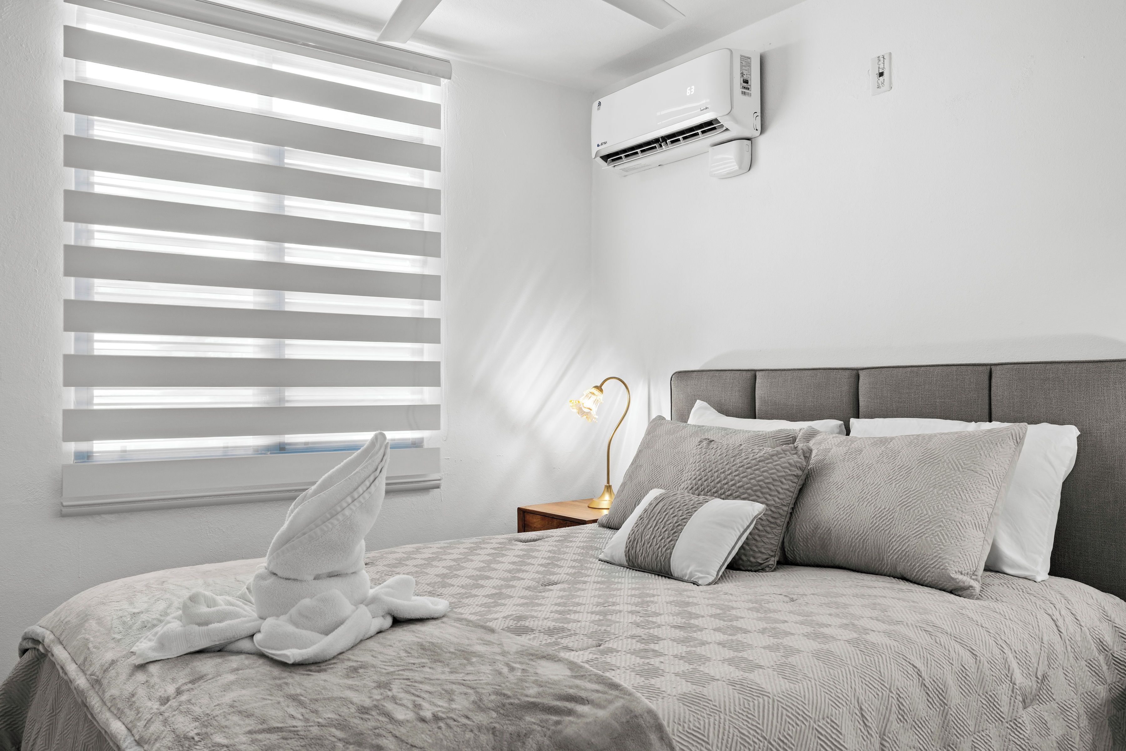 A bedroom with a bed, a bedside lamp, a window with blinds, and an air conditioning unit on the wall.
