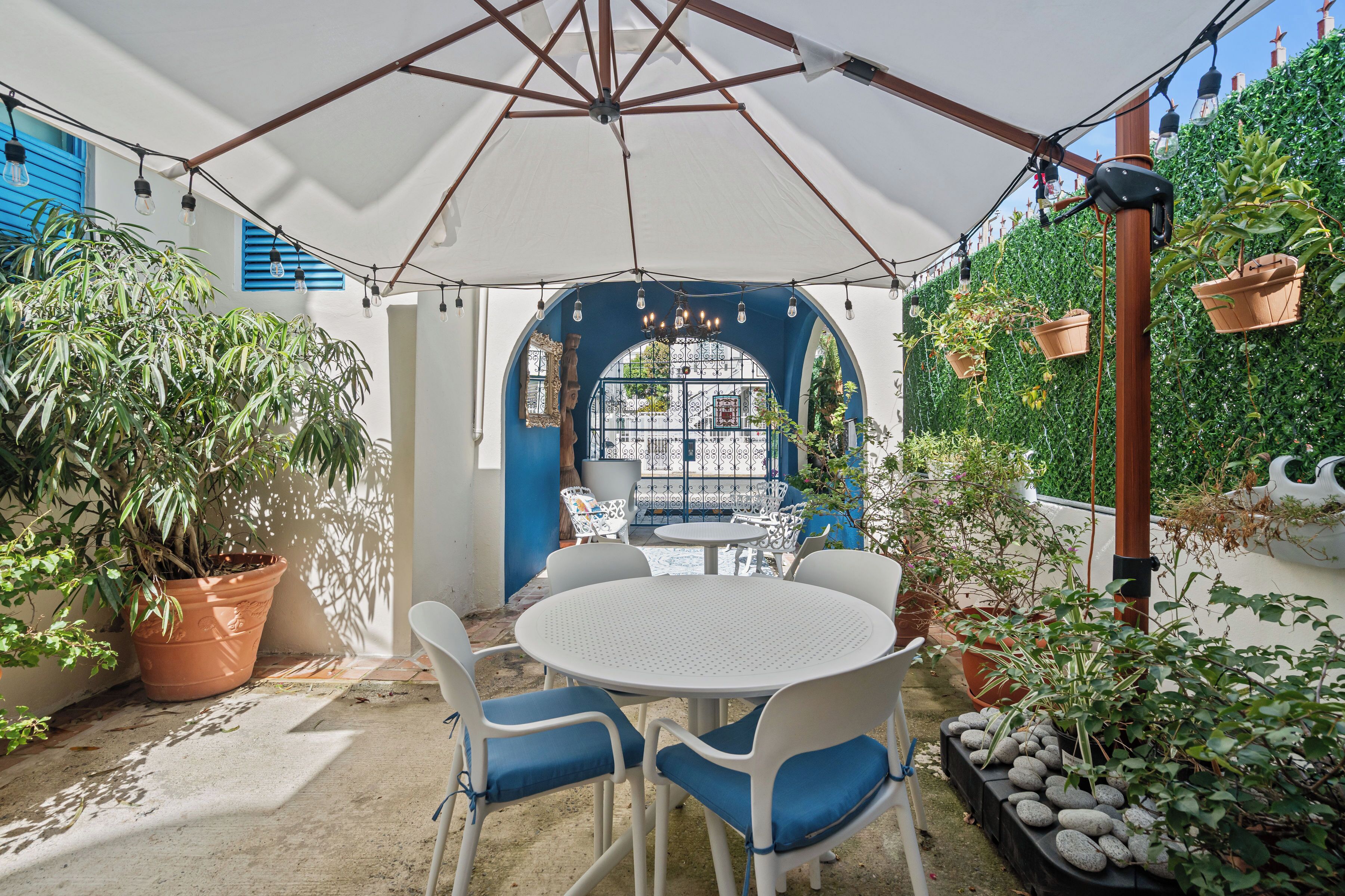 Een overdekt terras met een ronde tafel en stoelen, omgeven door plantenpotten en uitzicht op een blauw gebouw.