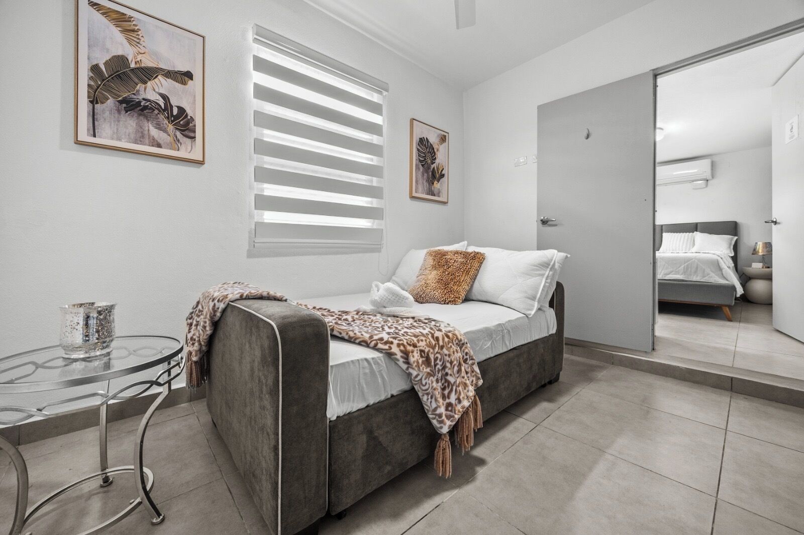 A modern bedroom with a grey sofa bed, a glass coffee table, and two framed artworks on the wall.