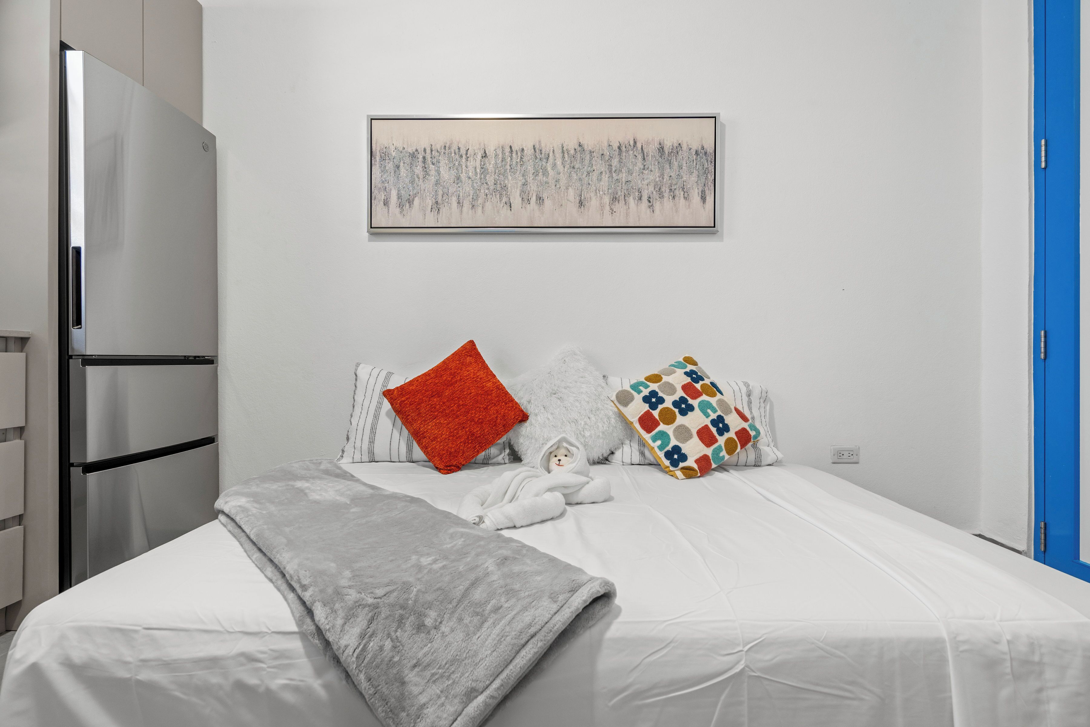 Un dormitorio con una cama, almohadas, una colcha, una obra de arte en la pared, un refrigerador y una puerta azul.