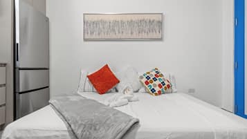 Un dormitorio con una cama, almohadas, una colcha, una obra de arte en la pared, un refrigerador y una puerta azul.