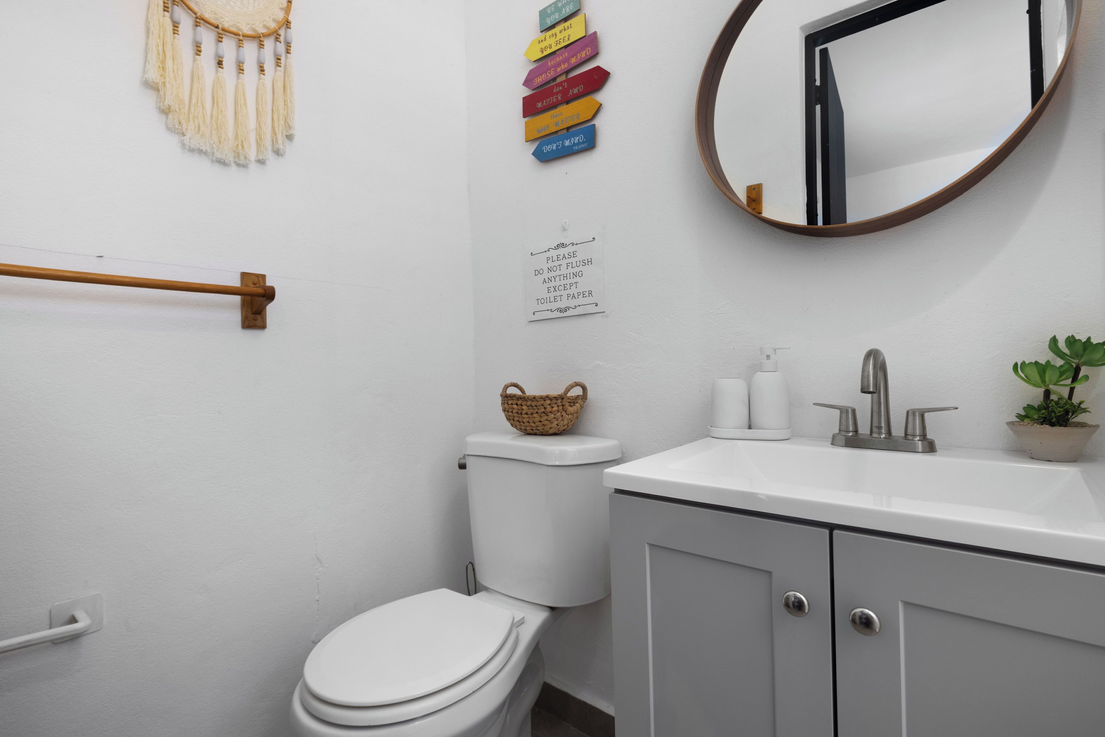 Un baño con inodoro, lavamanos, espejo, una cesta fijada a la pared y un cartel que prohíbe arrojar objetos que no sean papel.