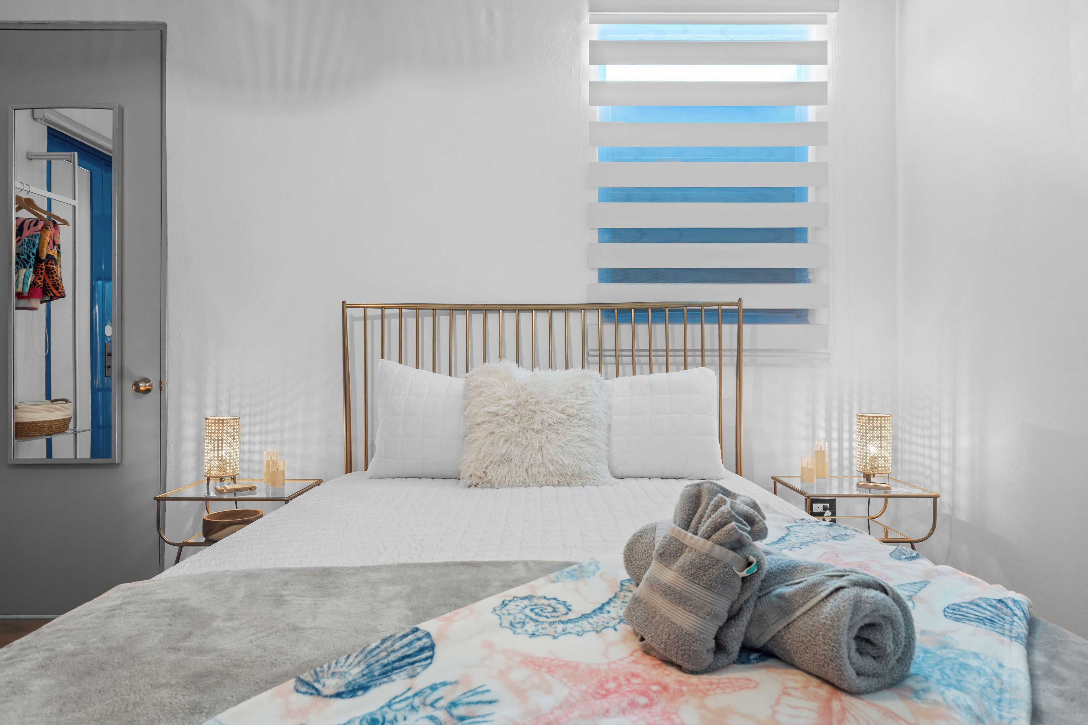 A hotel room with a bed, a window with blinds, a bedside table with a lamp, and a towel on the bed.