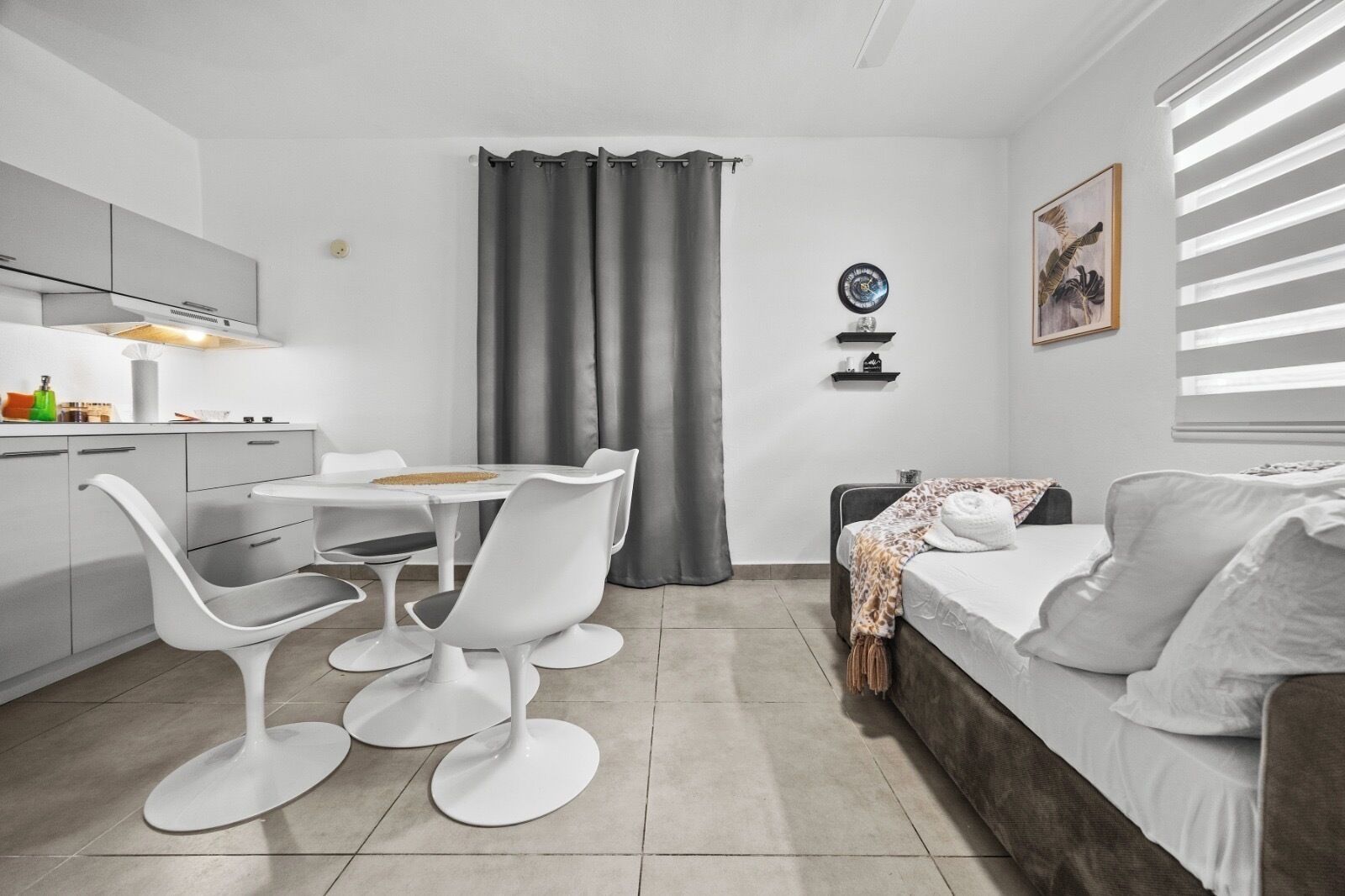 Una cocina moderna con armarios blancos, una mesa de comedor con sillas blancas, una cortina gris y una cama con almohada blanca.