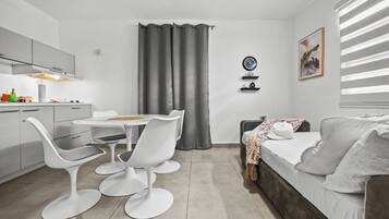 Una cocina moderna con armarios blancos, una mesa de comedor con sillas blancas, una cortina gris y una cama con almohada blanca.