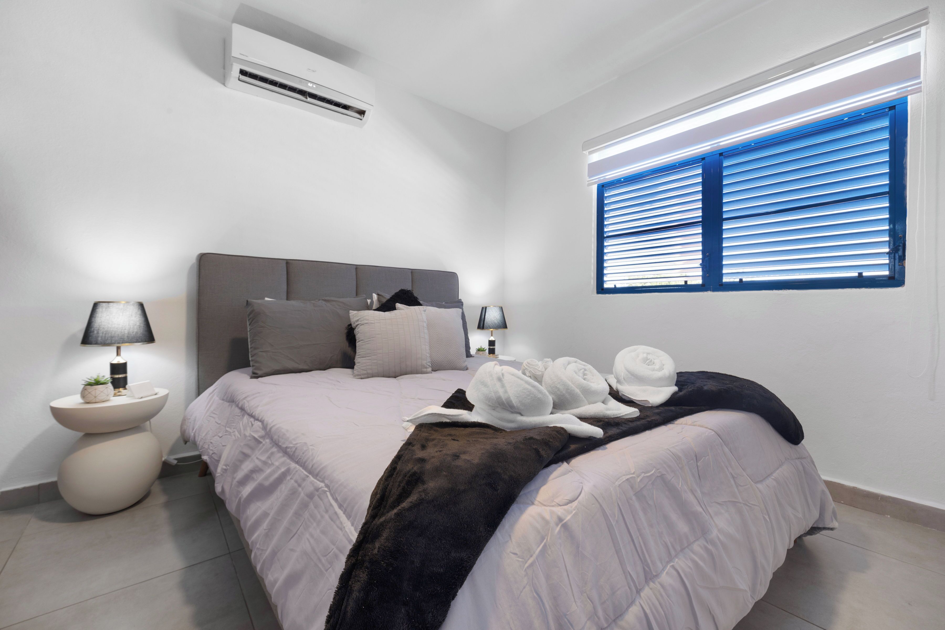 A modern bedroom with a bed, two bedside tables, a window with blinds, and an air conditioning unit.