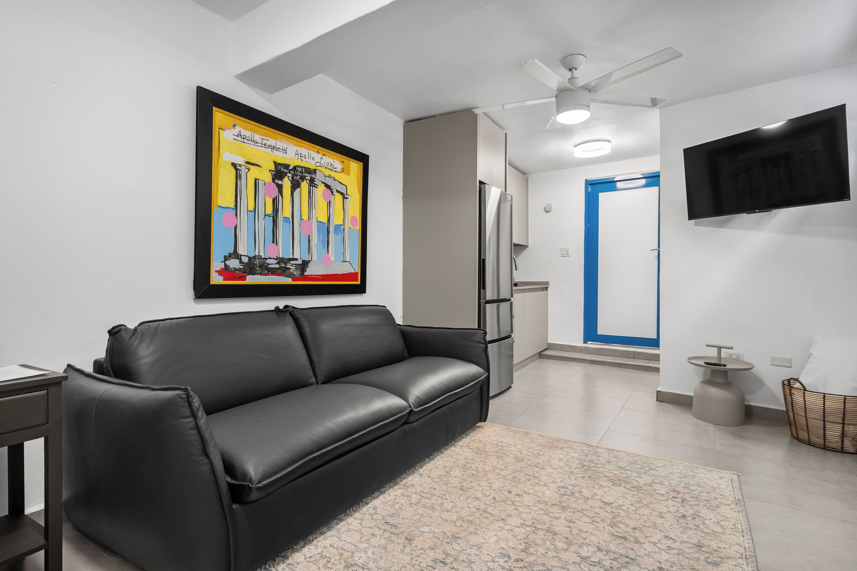 A modern living room with a black leather sofa, a ceiling fan, a flat-screen TV mounted on the wall, and a kitchen area with a refrigerator and cabinets.