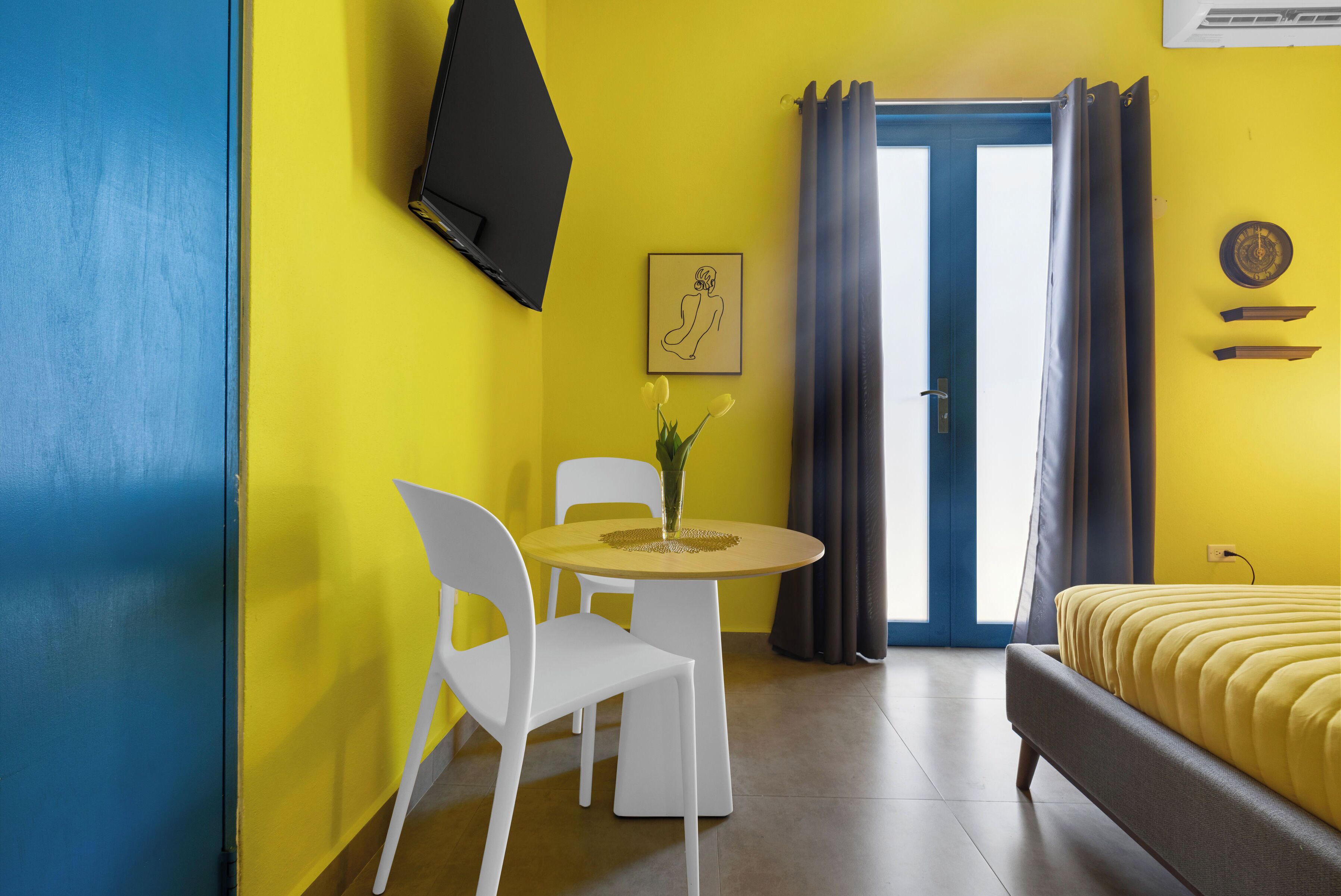 A room with a bed, a dining table with chairs, a TV mounted on the wall, and a window with curtains.