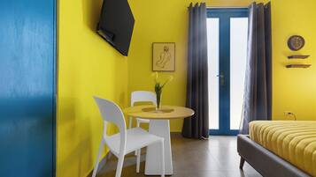 A room with a bed, a dining table with chairs, a TV mounted on the wall, and a window with curtains.