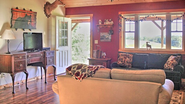 A cozy living room with a wooden ceiling, a television on a wooden stand, a sofa with patterned cushions, and a large window with a view of a dog and greenery outside. - Picturesque Cabin set on 150-acre farm, 5-minute drive to Chautauqua Institute (Mayville)