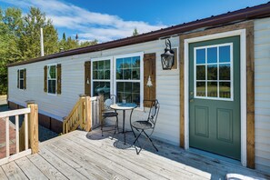 Una terraza de madera con una mesa y sillas, una puerta verde y una ventana con vistas a árboles.